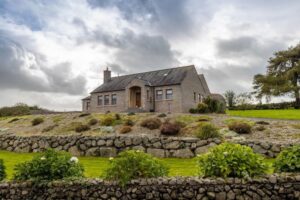 The front exterior of 53 Bettys Hill Road. The is a detached bungalow for sale in Ballyholland. Countryside surrounds this property. Well maintained gardens can be seen which contains many plants and shrubs. There is also stone walls.
