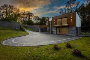 The front exterior of 37 Glen Road. This is a contemporary detached property for sale in Cultra. The exterior of the property is a mix of stone, render, glass and treated hardwood. A large tiled area leads to the front of the property, offering parking space for multiple vehicles.